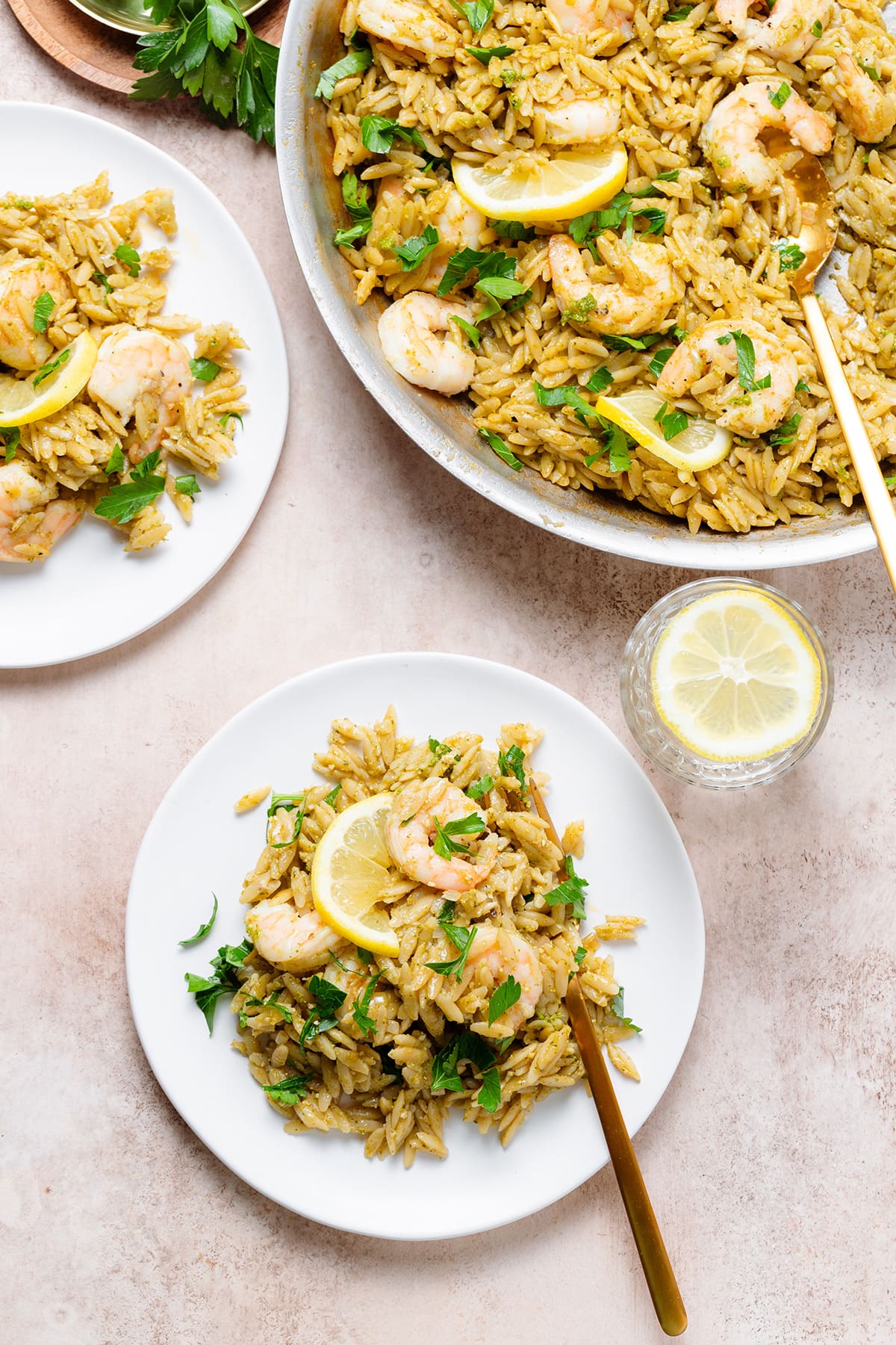 Pesto orzo with shrimp and a slice of lemon on a white plate with more orzo in a pan in the top right corner.