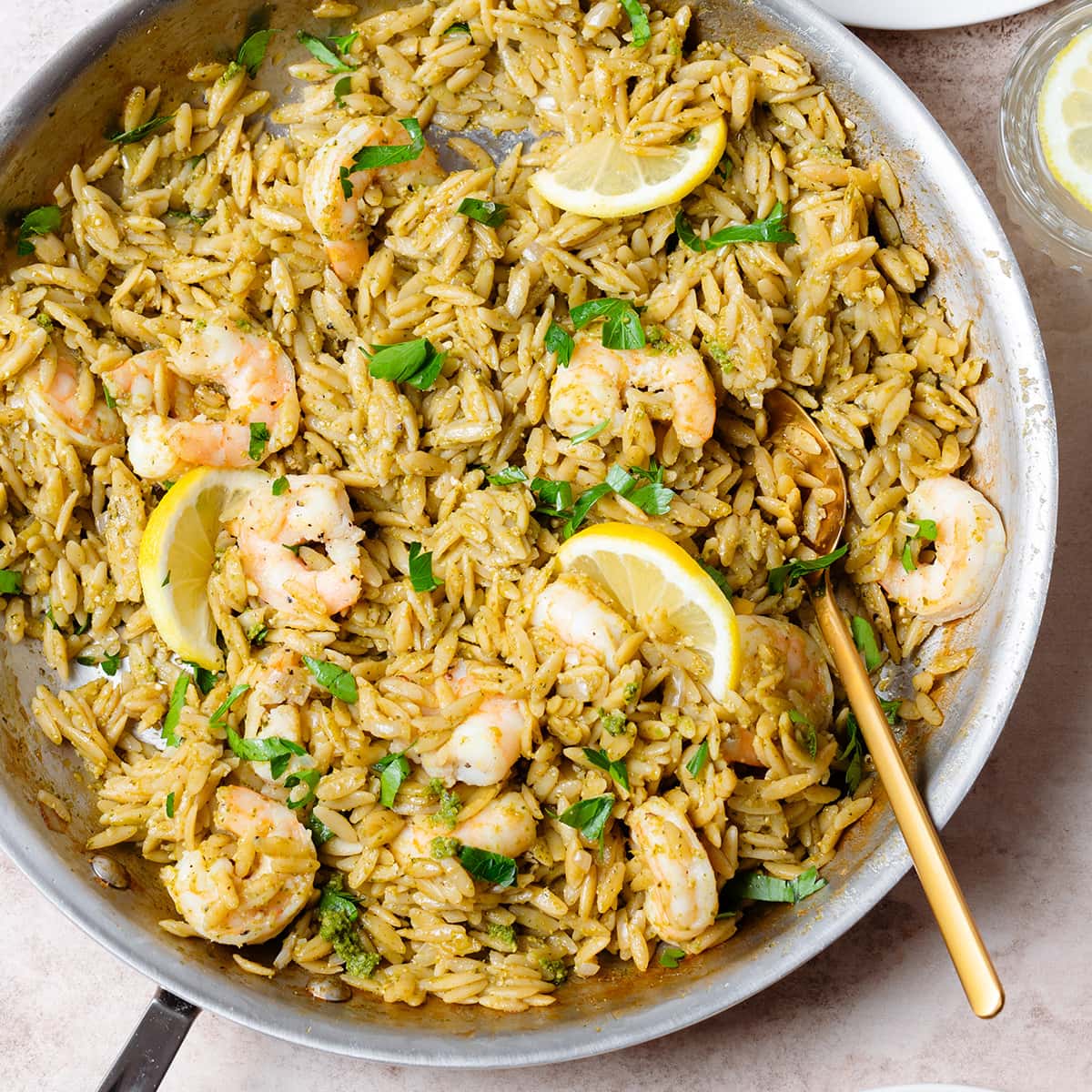 Pesto orzo with shrimp and slices of lemon in a large pan with a gold spoon resting on the right side of it.