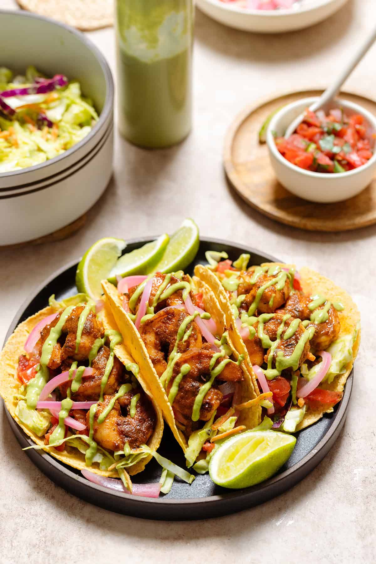 Three corn tortilla tacos with shrimp, slaw, green sauce, and lime wedges on a black plate.