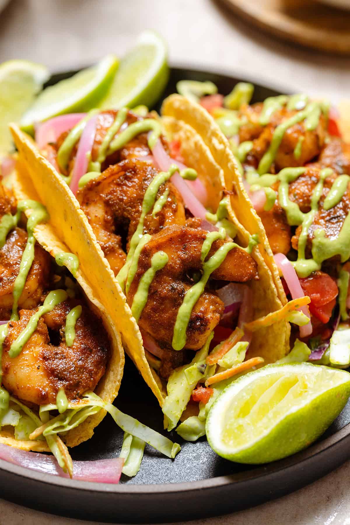 Three corn tortilla tacos with shrimp, slaw, green sauce on a black plate.