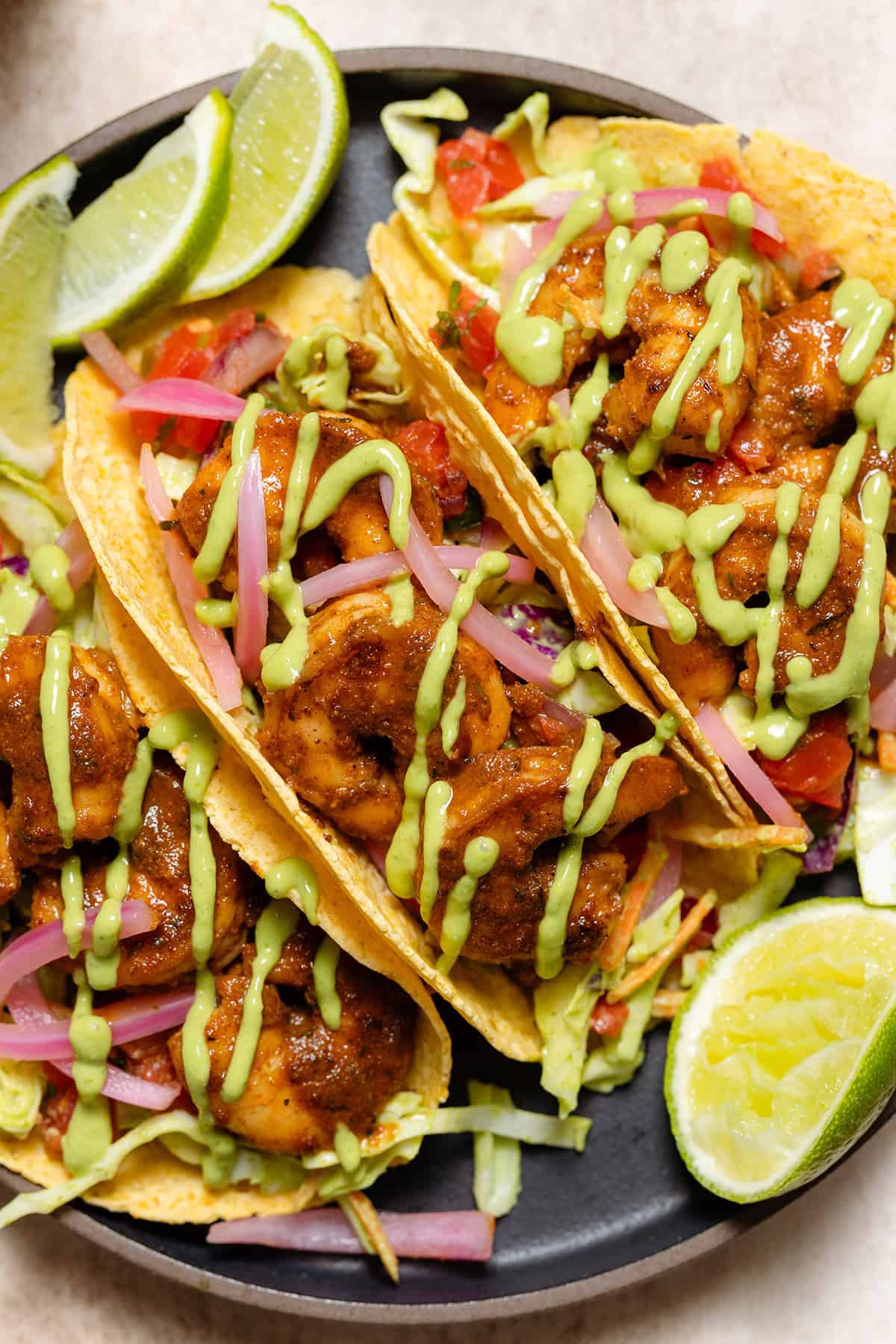 Three corn tortilla tacos with shrimp, slaw, green sauce, and lime wedges on a black plate.