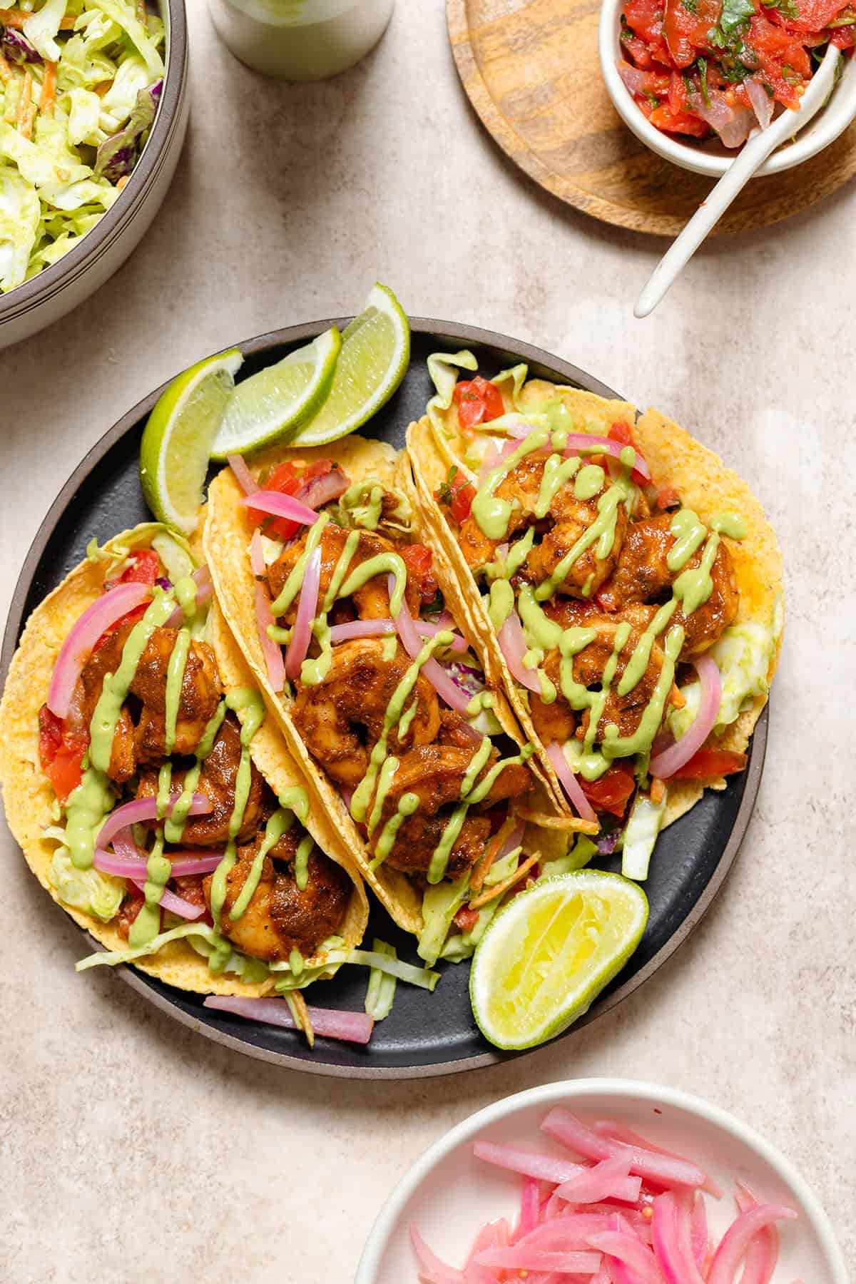 Three corn tortilla tacos with shrimp, slaw, green sauce, and lime wedges on a black plate.