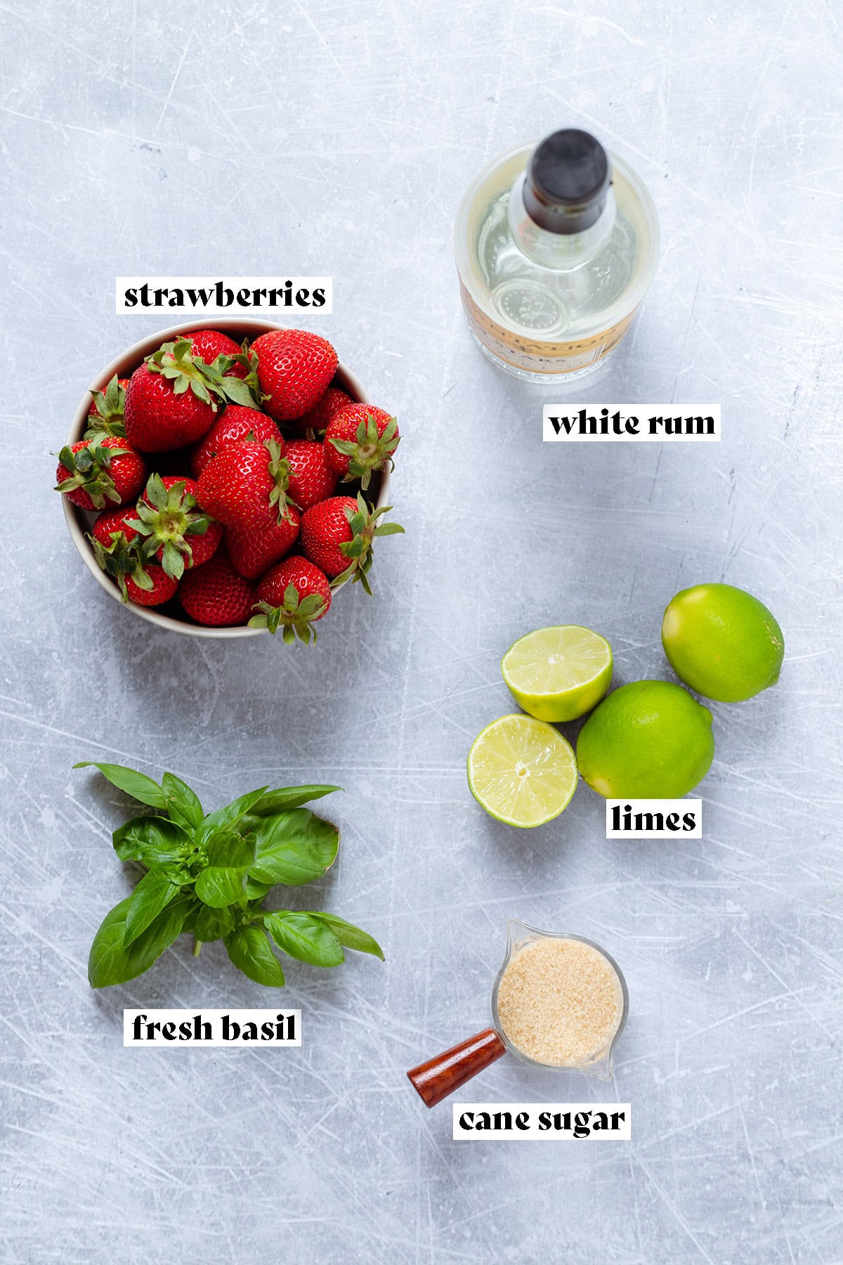 Ingredients like fresh strawberries, lime, basil, and rum laid out on a metal background.