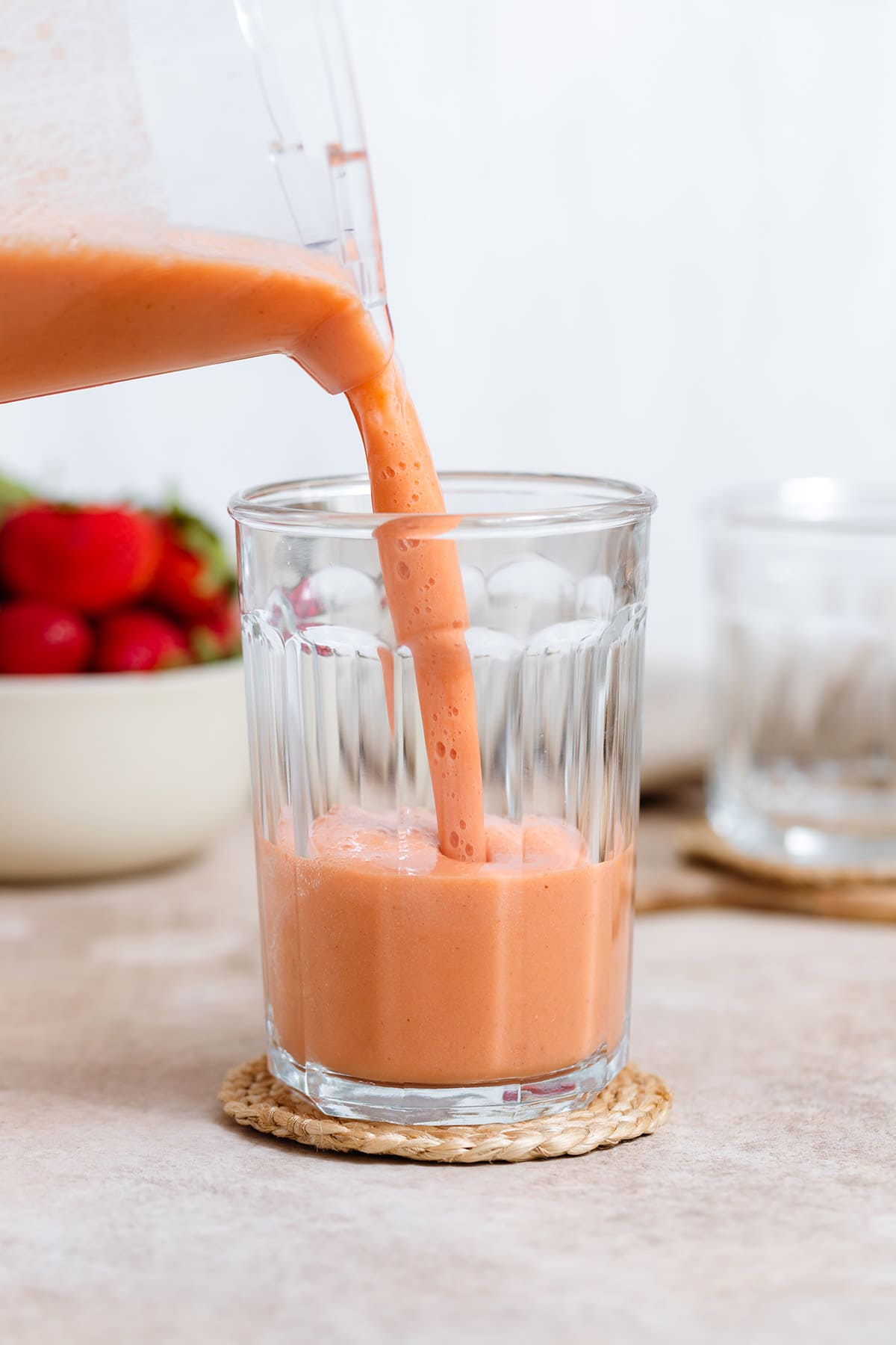 Strawberry peach smoothie being poured into a tall glass on a beige background.