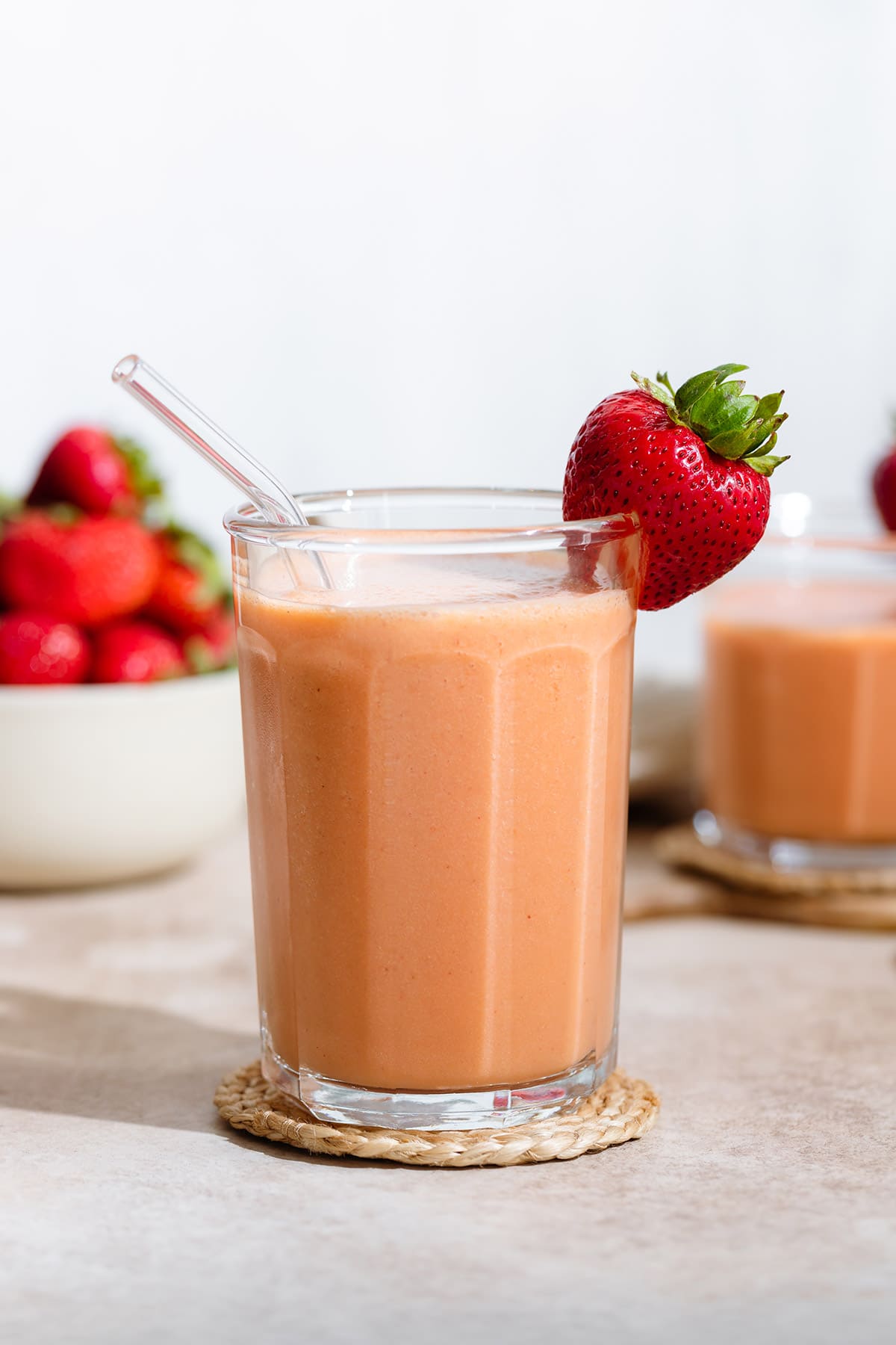Peach colored smoothie in a tall glass with a glass straw and garnished with a fresh strawberry on the rim and more strawberries around it.
