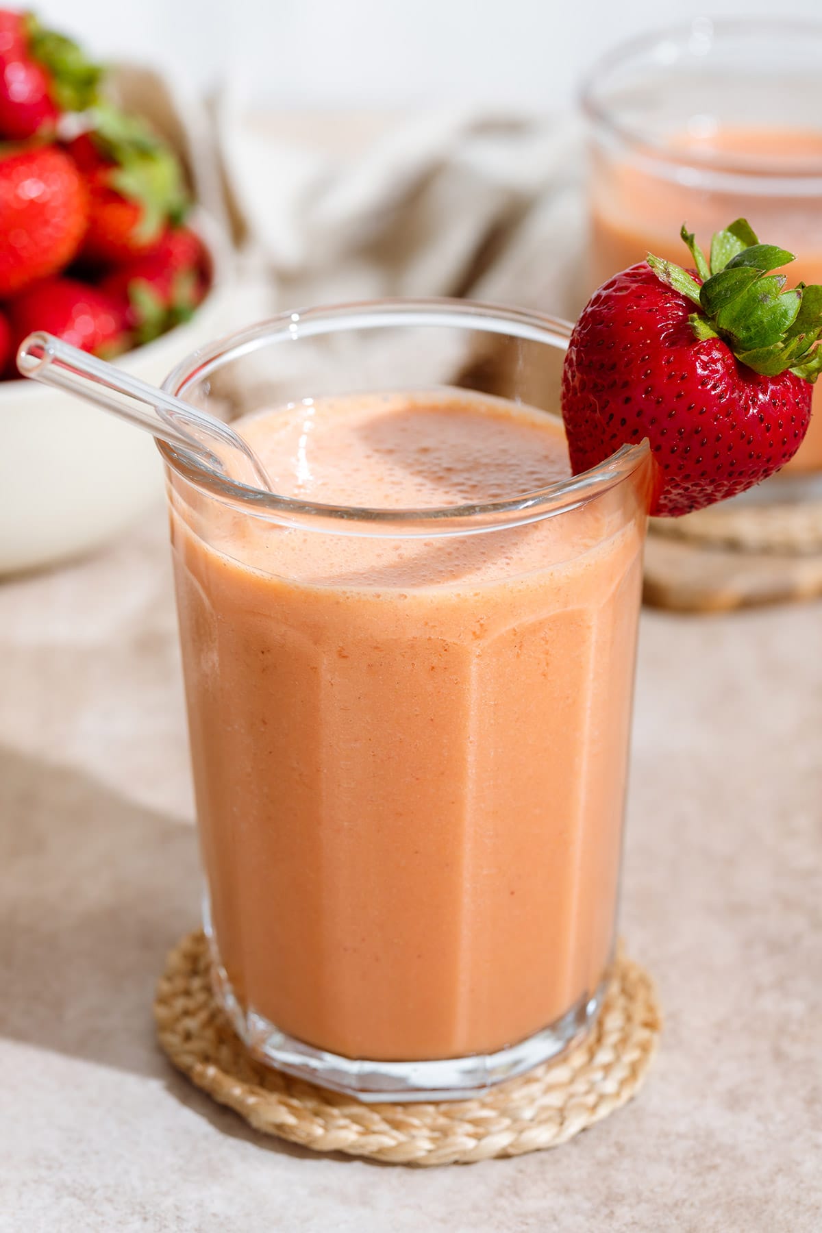 Peach colored smoothie in a tall glass with a glass straw and garnished with a fresh strawberry on the rim and more strawberries around it.