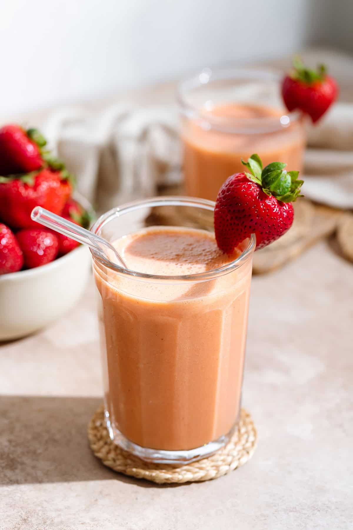 Peach strawberry smoothie in a tall glass with a glass straw and garnished with a fresh strawberry on the rim and more strawberries around it.