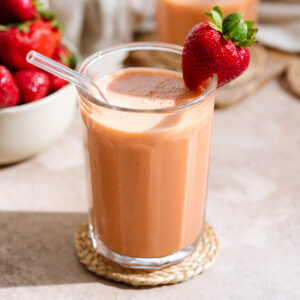 Peach colored smoothie in a tall glass with a glass straw and garnished with a fresh strawberry on the rim.