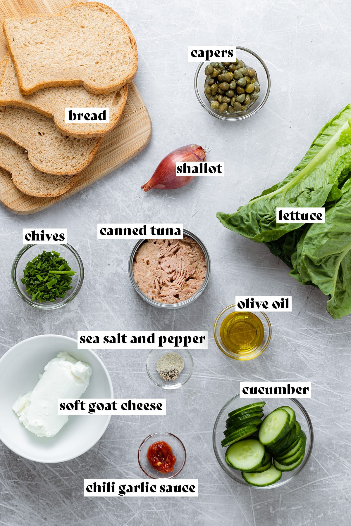 Ingredients for tuna sandwich like bread, canned tuna, and sliced cucumber laid out on a metal background.