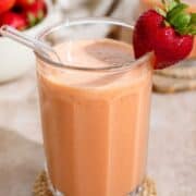 Peach colored smoothie in a tall glass with a glass straw and garnished with a fresh strawberry on the rim and more strawberries around it.