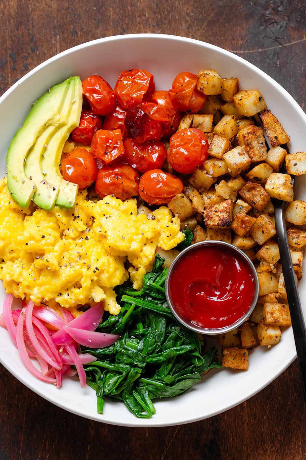 A bowl with scrambled eggs, roasted potatoes, roasted tomatoes, spinach, and ketchup.