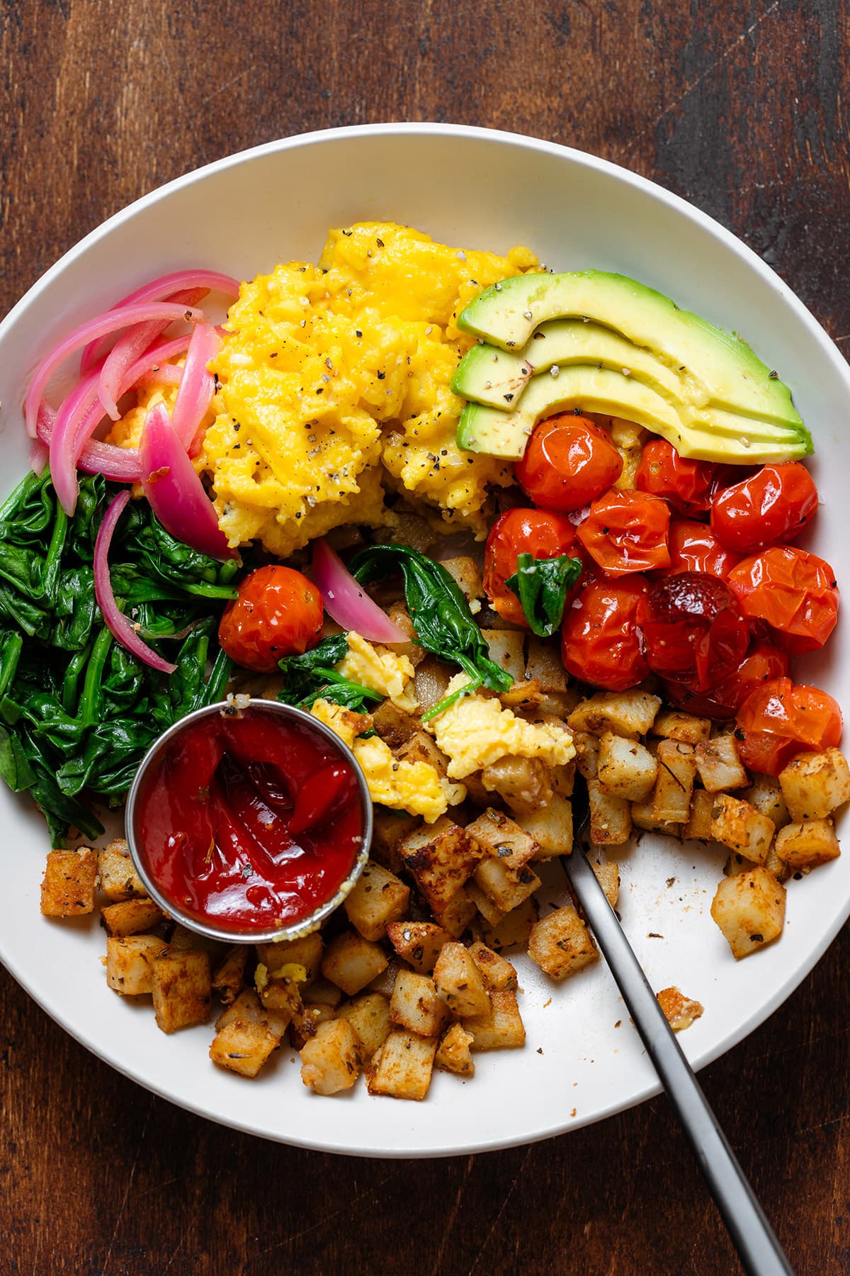 A bowl with scrambled eggs, roasted potatoes, roasted tomatoes, spinach, and ketchup, partially eaten.