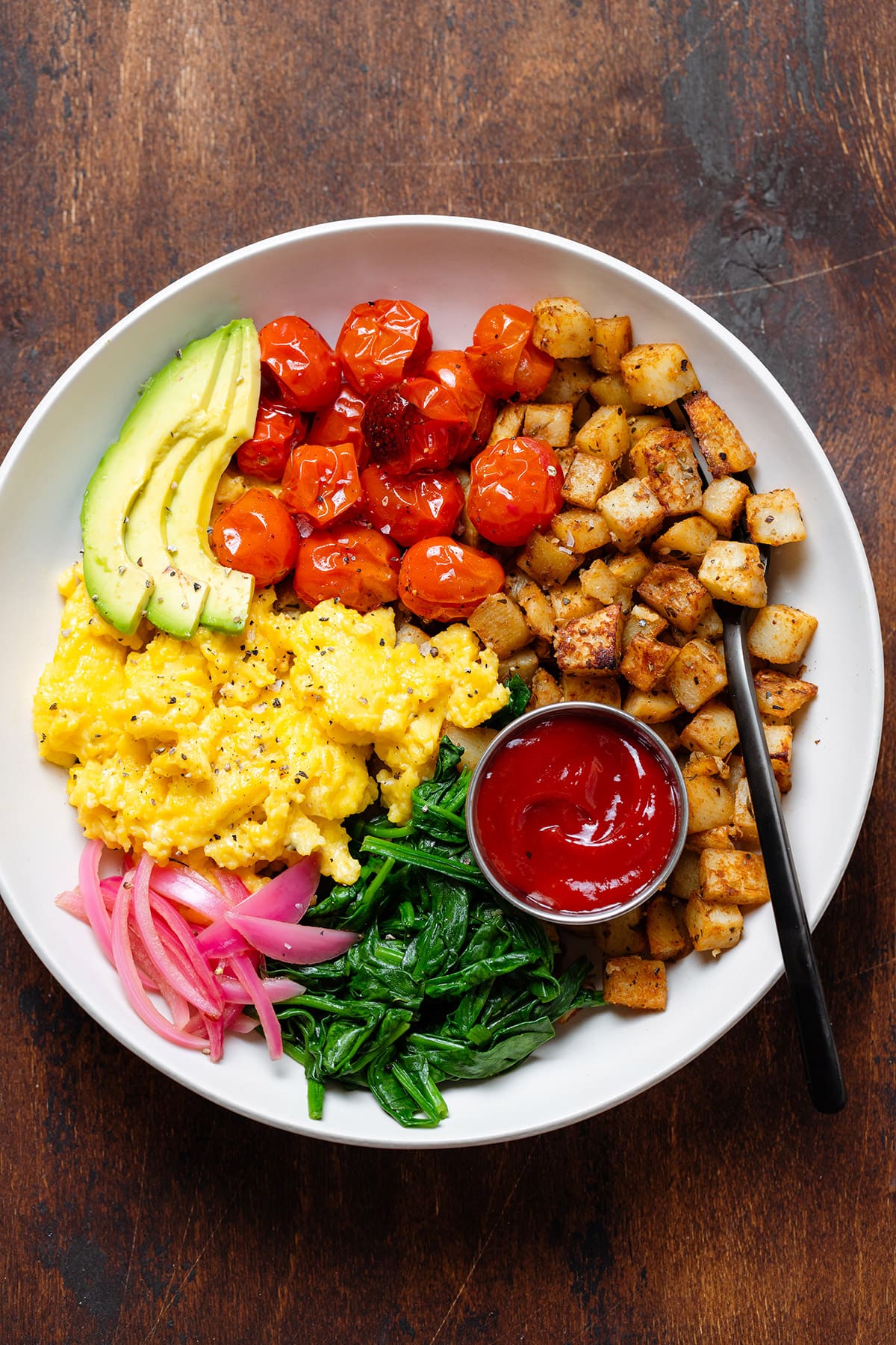 A bowl with scrambled eggs, roasted potatoes, roasted tomatoes, spinach, and ketchup.