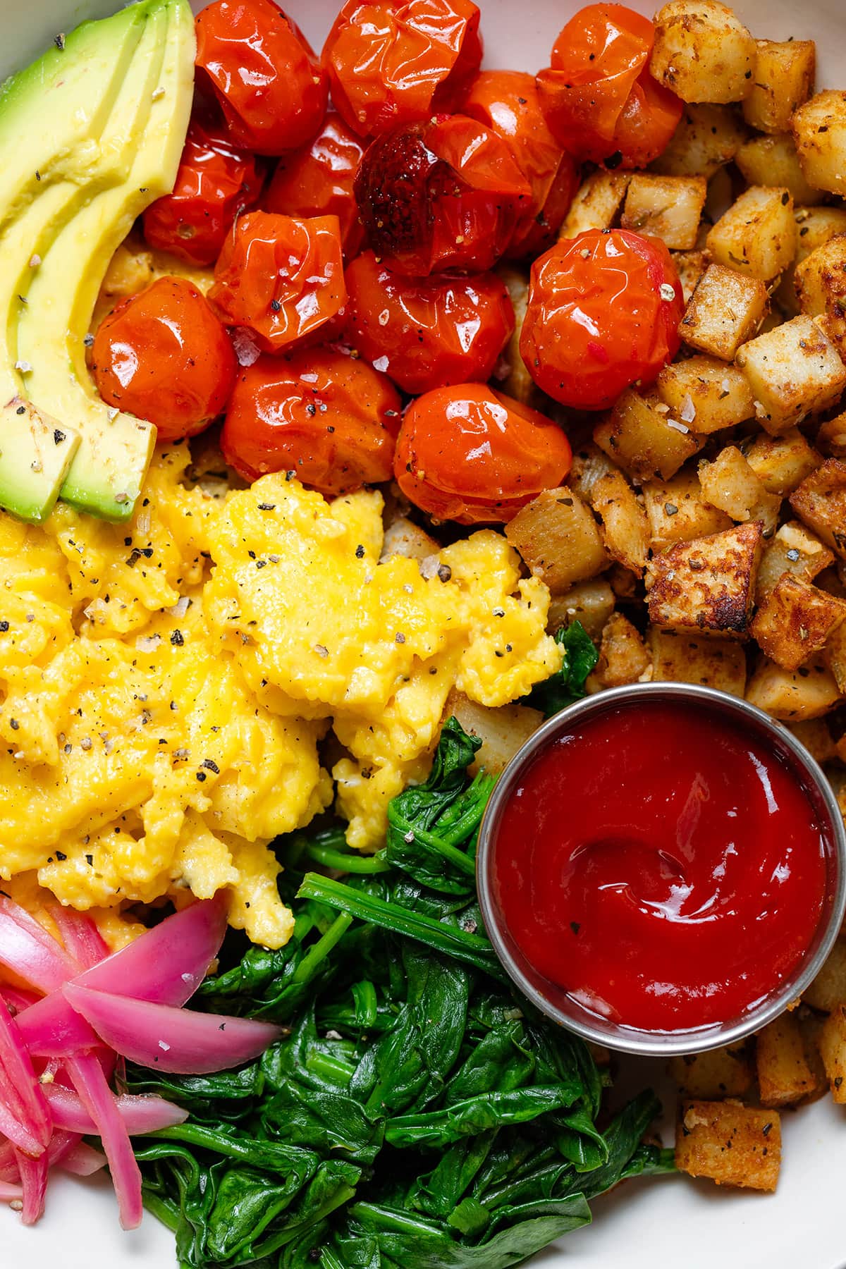 A bowl with scrambled eggs, roasted potatoes, roasted tomatoes, spinach, and ketchup.