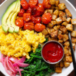 A bowl with scrambled eggs, roasted potatoes, roasted tomatoes, spinach, and ketchup.