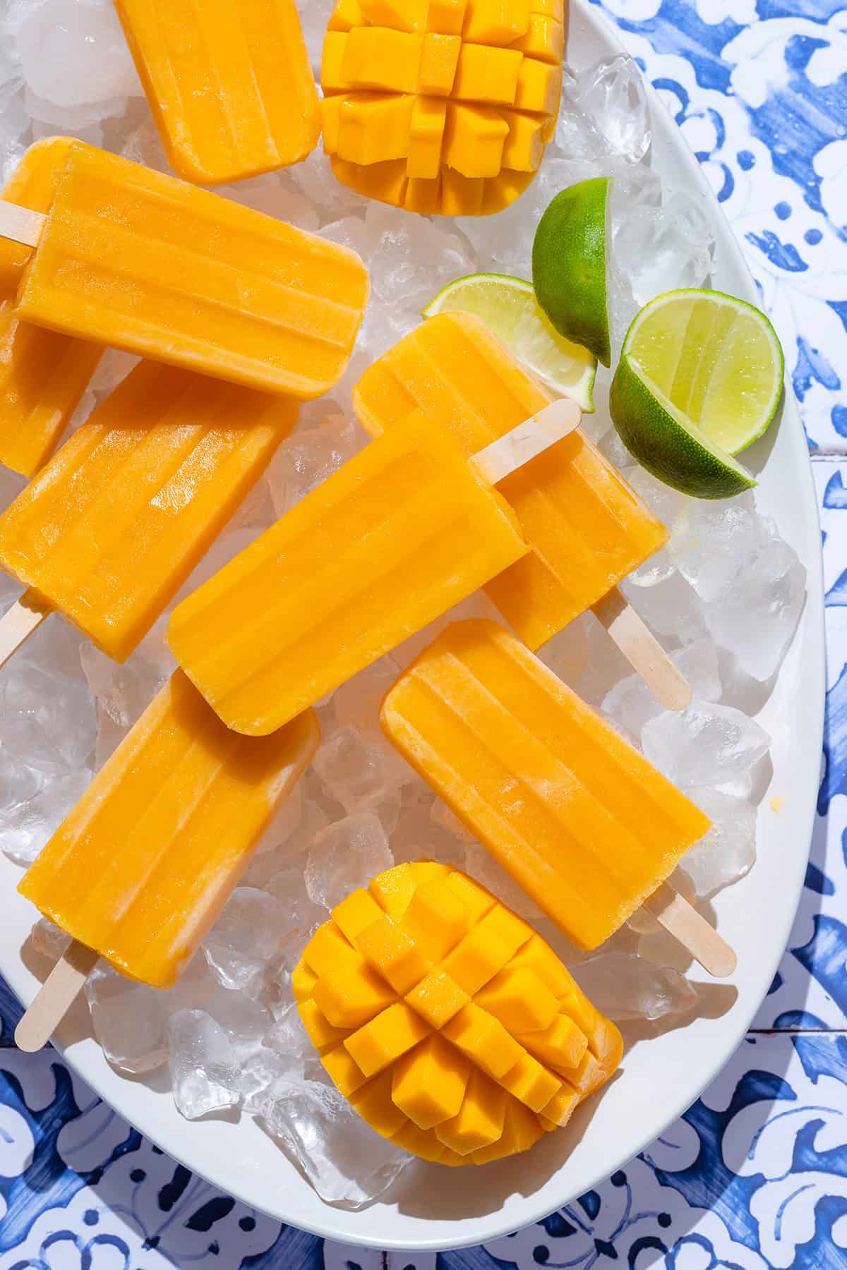 Bright orange mango popsicles on a white serving plate with ice and lime slices on a blue tile background.