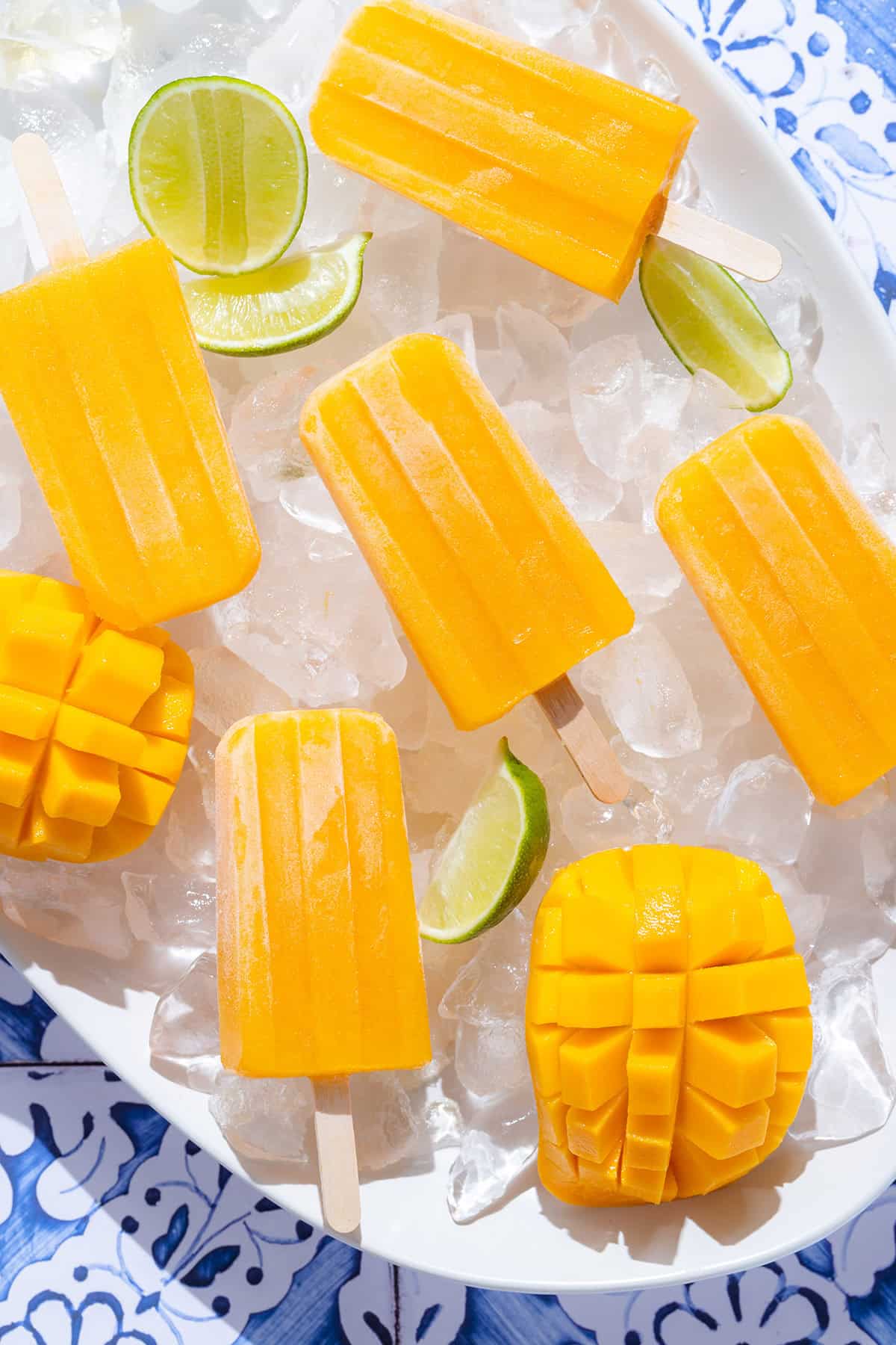 Bright orange mango popsicles on a white serving plate with ice and lime slices on a blue tile background.