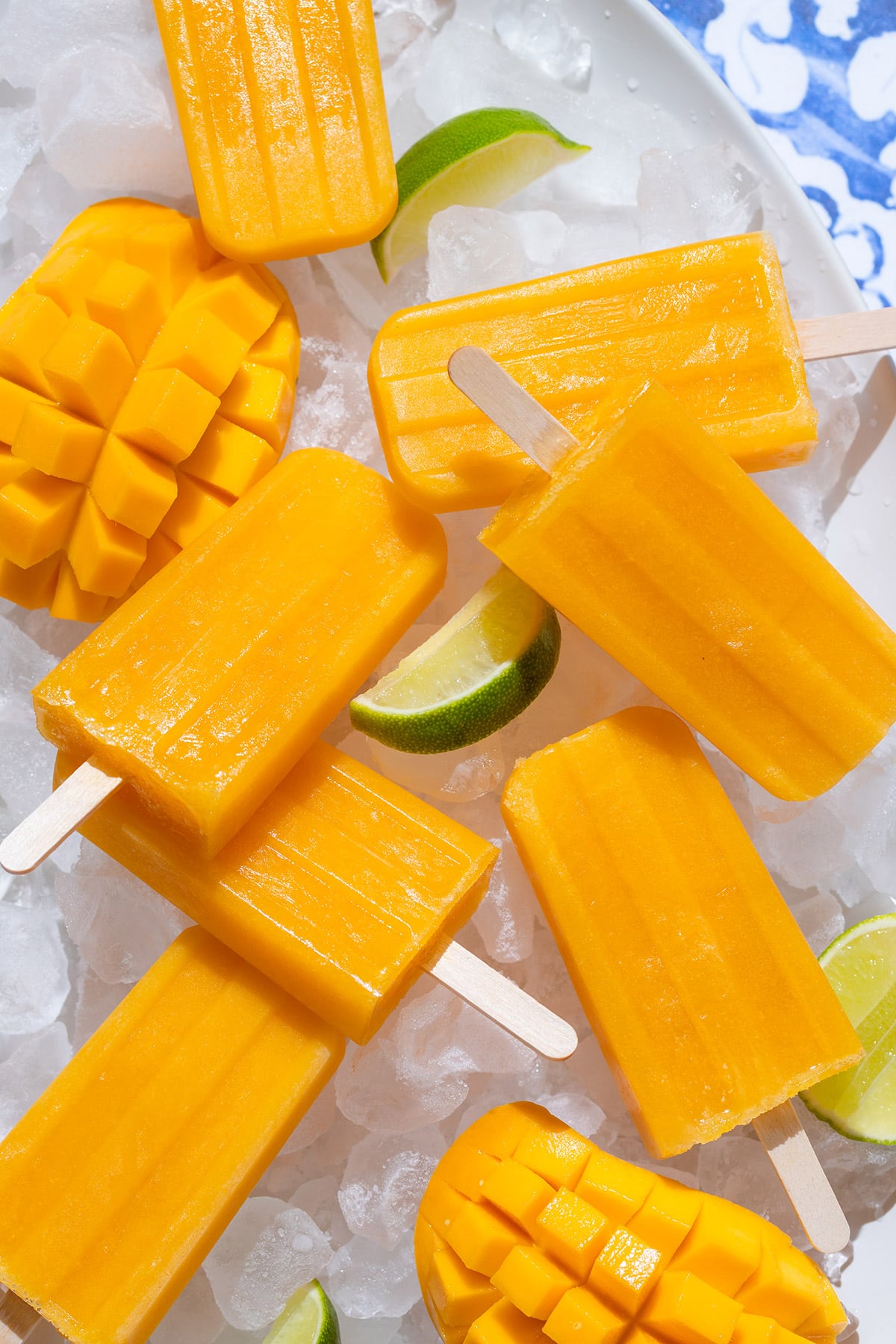 Bright orange mango popsicles on a white serving plate with ice and lime slices on a blue tile background.
