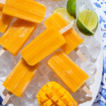Bright orange mango popsicles on a white serving plate with ice and lime slices.