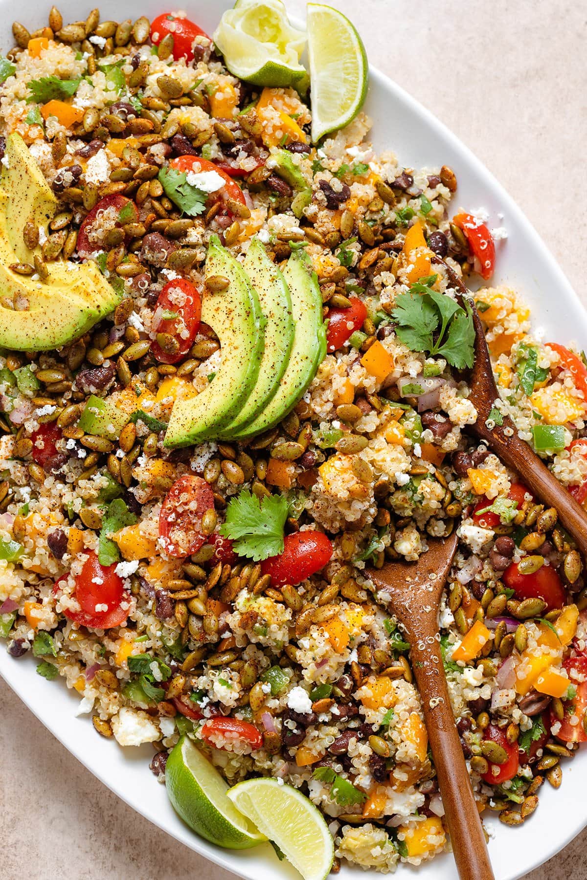 Mango quinoa salad topped with avocado slices on a large serving plate with wooden serving spoons on the right side.