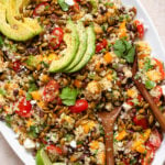 Mango quinoa salad topped with avocado slices on a large serving plate with wooden serving spoons on the right side.