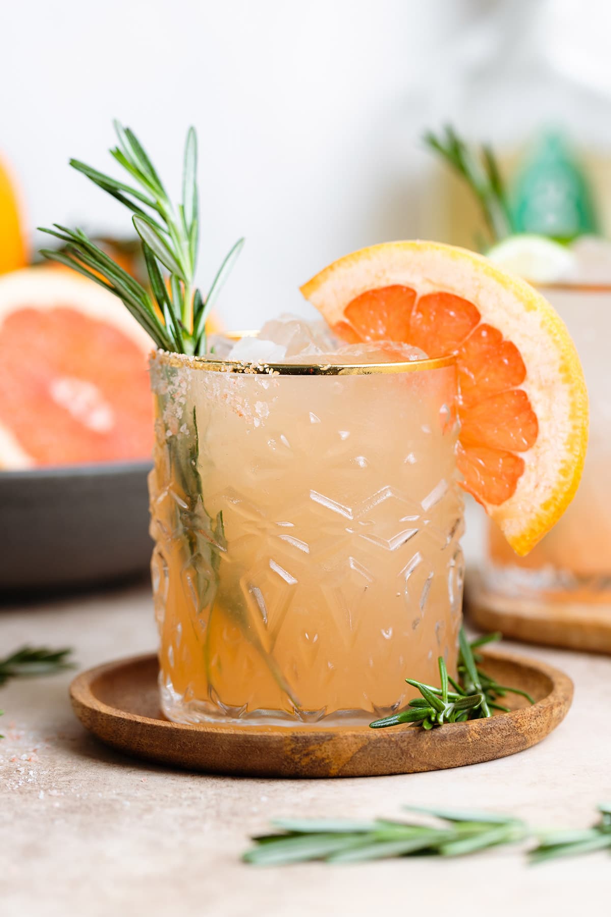 Rosemary paloma cocktail in a short glass with a gold rim garnished with fresh rosemary and a grapefruit slice.
