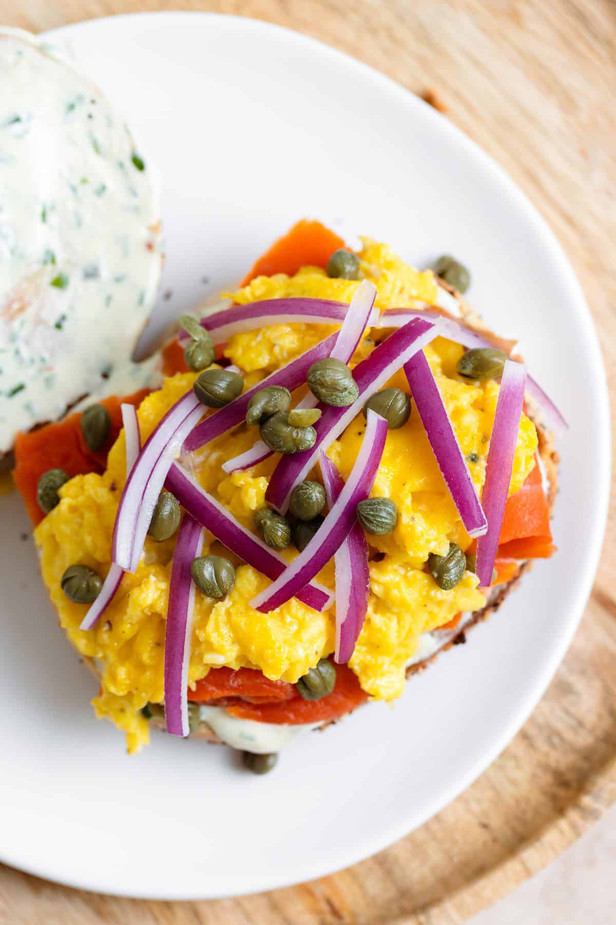 Open face toasted everything bagel with smoked salmon, scrambled eggs, cream cheese, capers, and red onion on a white plate.