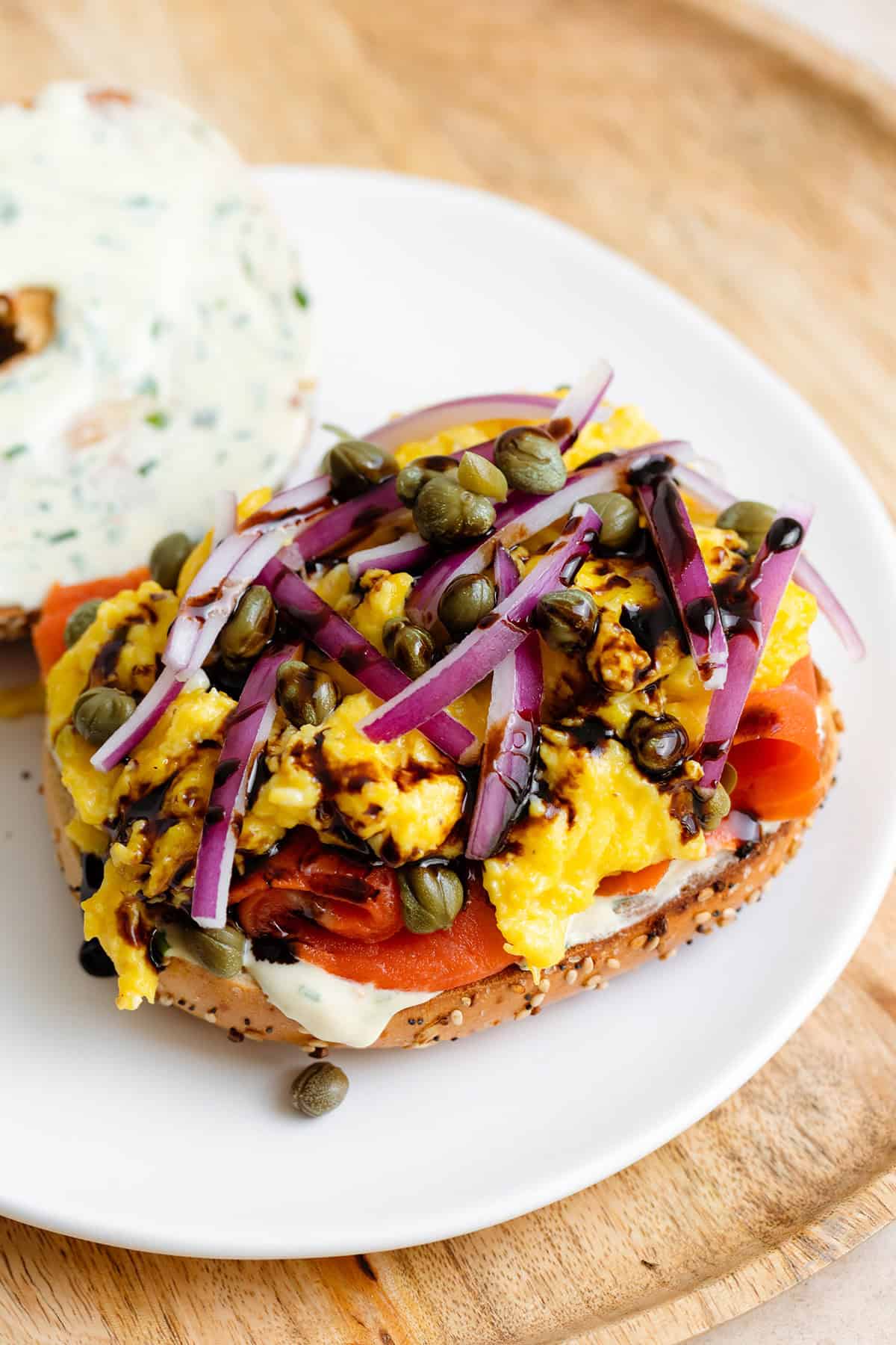 Open face toasted everything bagel with smoked salmon, scrambled eggs, cream cheese, capers, and red onion on a white plate.