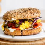 A toasted everything bagel with smoked salmon, scrambled eggs, cream cheese, capers, and red onion on a white plate.