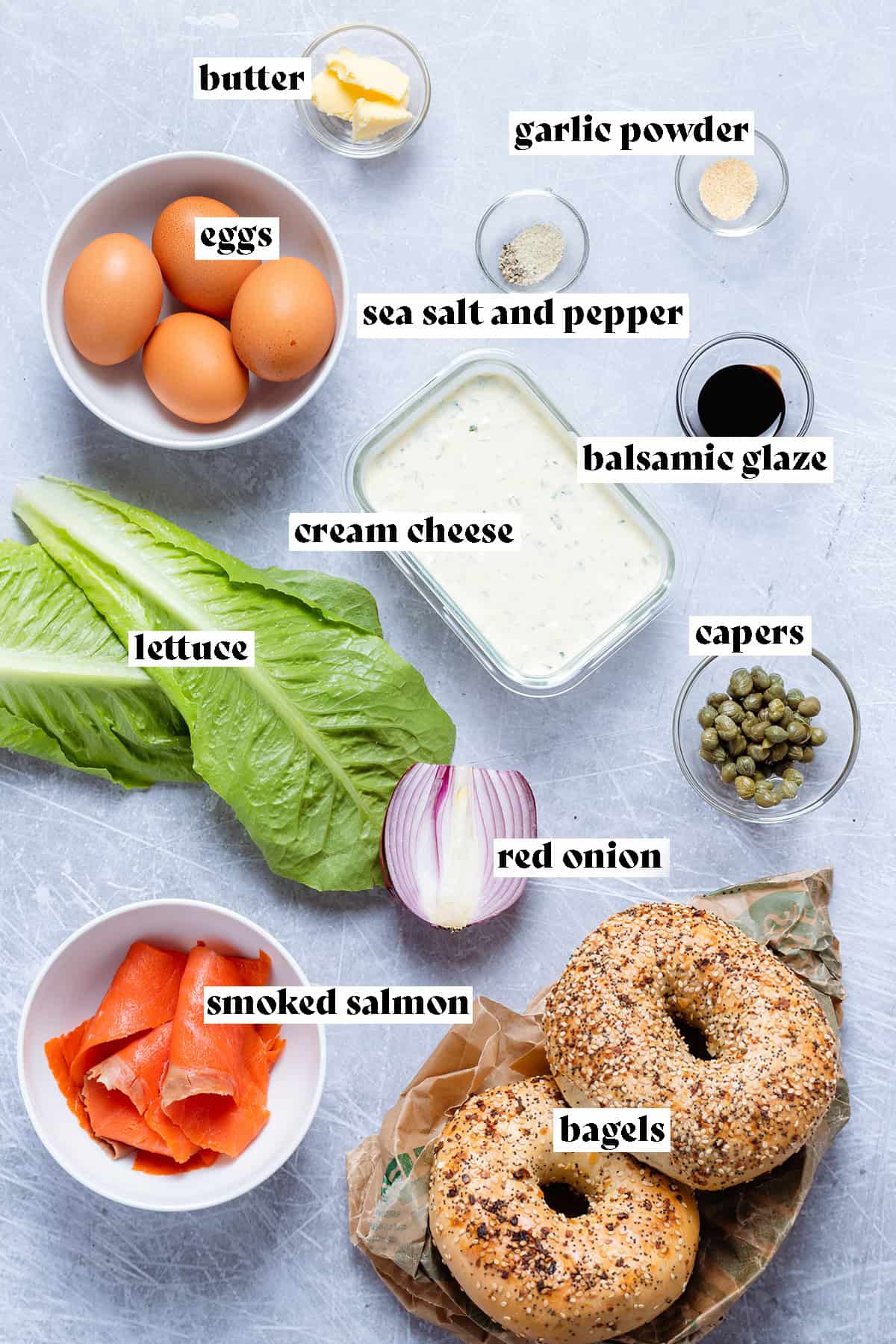 Ingredients like bagels, eggs, and cream cheese for a smoked salmon egg bagel all laid out on a grey background.