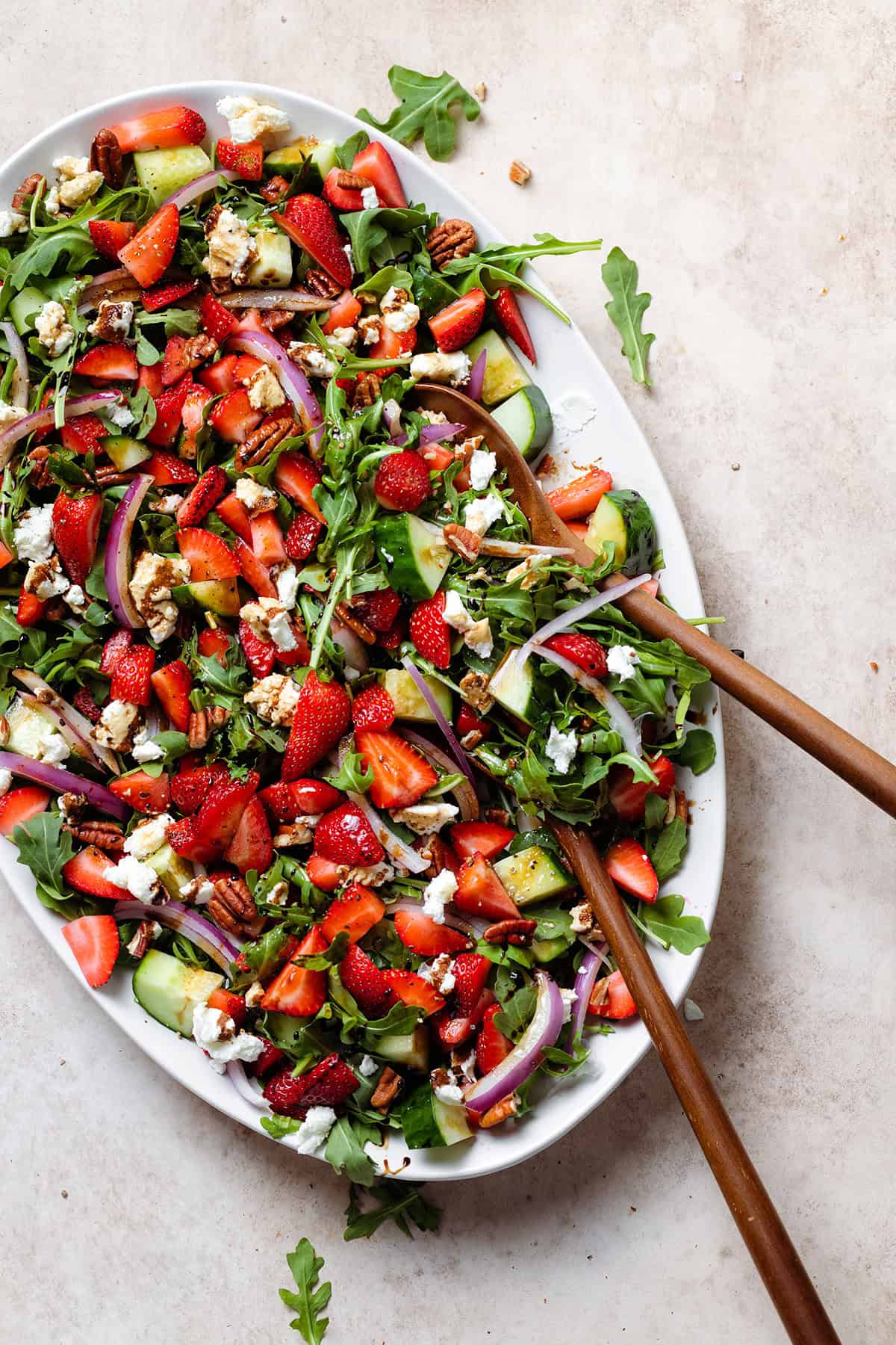 Strawberry goat cheese salad drizzled with balsamic glaze on a large white plate with wooden serving spoons.