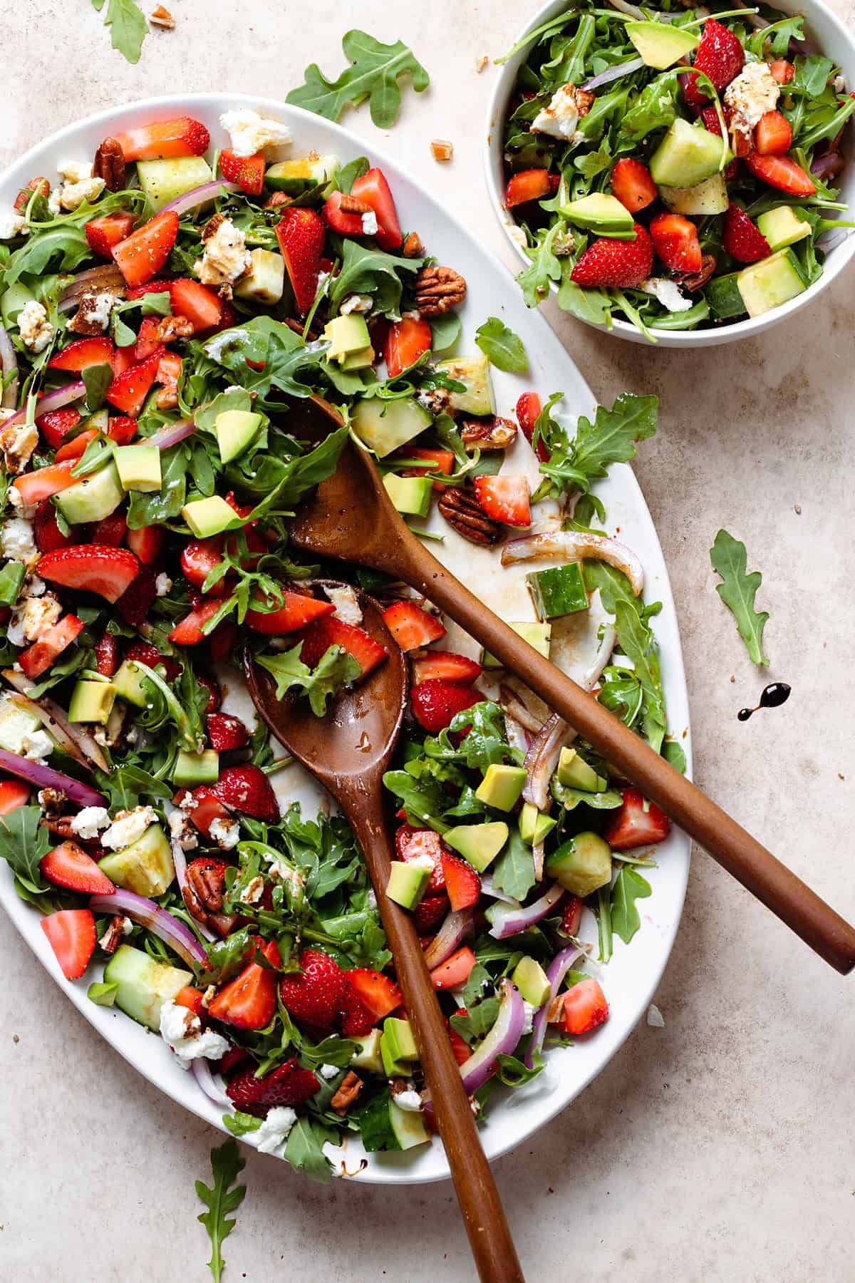 Strawberry goat cheese salad drizzled with balsamic glaze on a large white plate with wooden serving spoons.