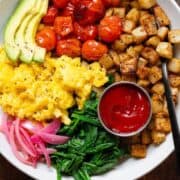 A bowl with scrambled eggs, roasted potatoes, roasted tomatoes, spinach, and ketchup.