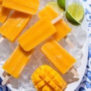 Bright orange mango popsicles on a white serving plate with ice and lime slices on a blue tile background.