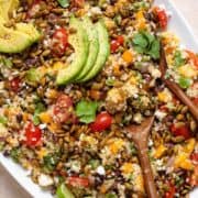 Mango quinoa salad topped with avocado slices on a large serving plate with wooden serving spoons on the right side.