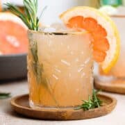 Rosemary paloma cocktail in a short glass with a gold rim garnished with fresh rosemary and a grapefruit slice.