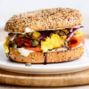 A toasted everything bagel with smoked salmon, scrambled eggs, cream cheese, capers, and red onion on a white plate.