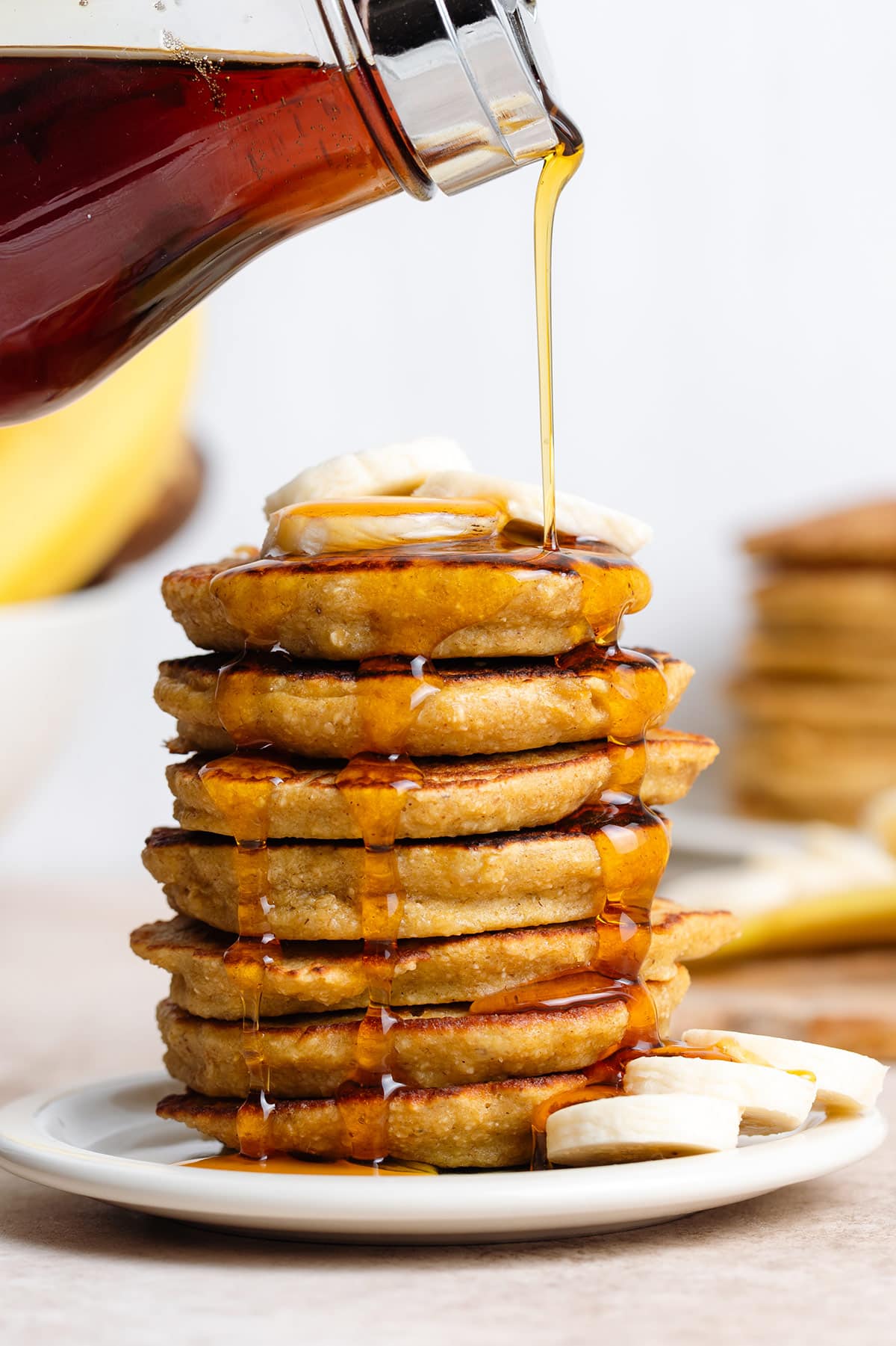 Maple syrup being poured over a stack of pancakes with banana slices on top.