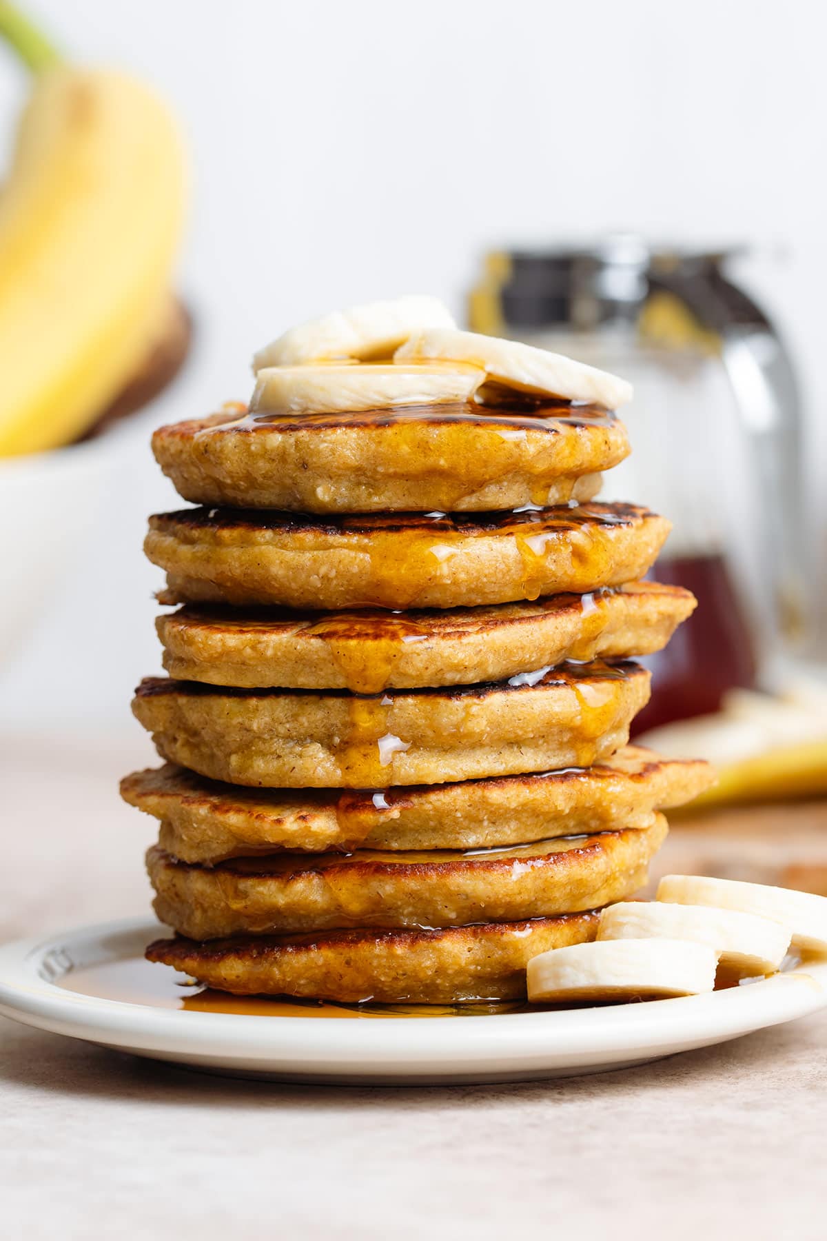Pancake stack topped with banana slices on a small white plate with bananas and maple syrup in the background.