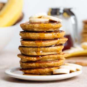 Pancake stack topped with banana slices on a small white plate with bananas and maple syrup in the background.