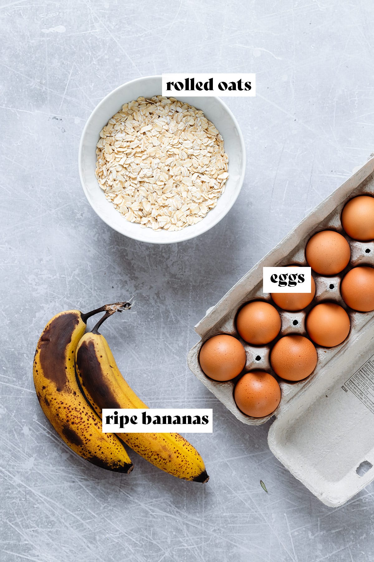 Two bananas, rolled oats in a bowl, and eggs in an egg carton on a grey background.