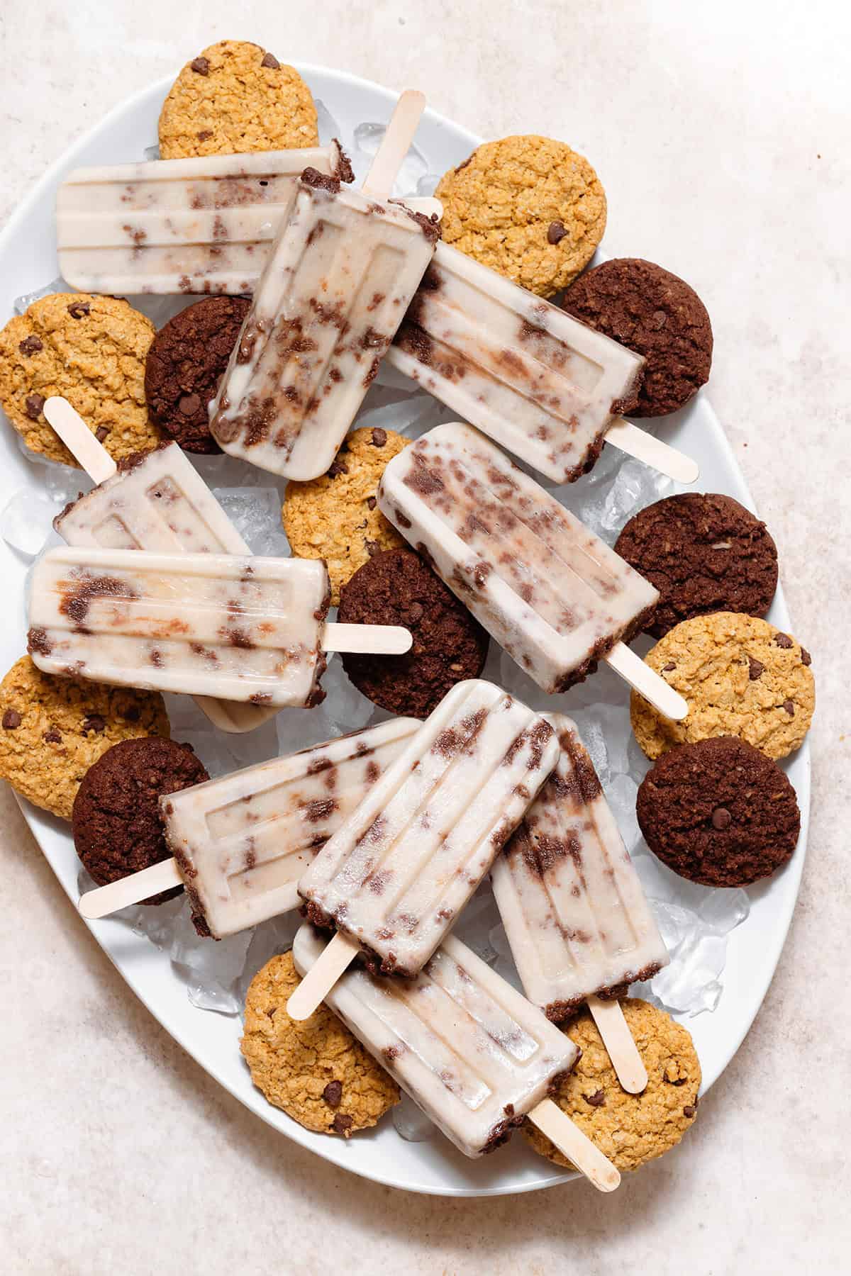 Cookie-dough popsicles arranged on a white serving platter filled with ice and chocolate cookies.