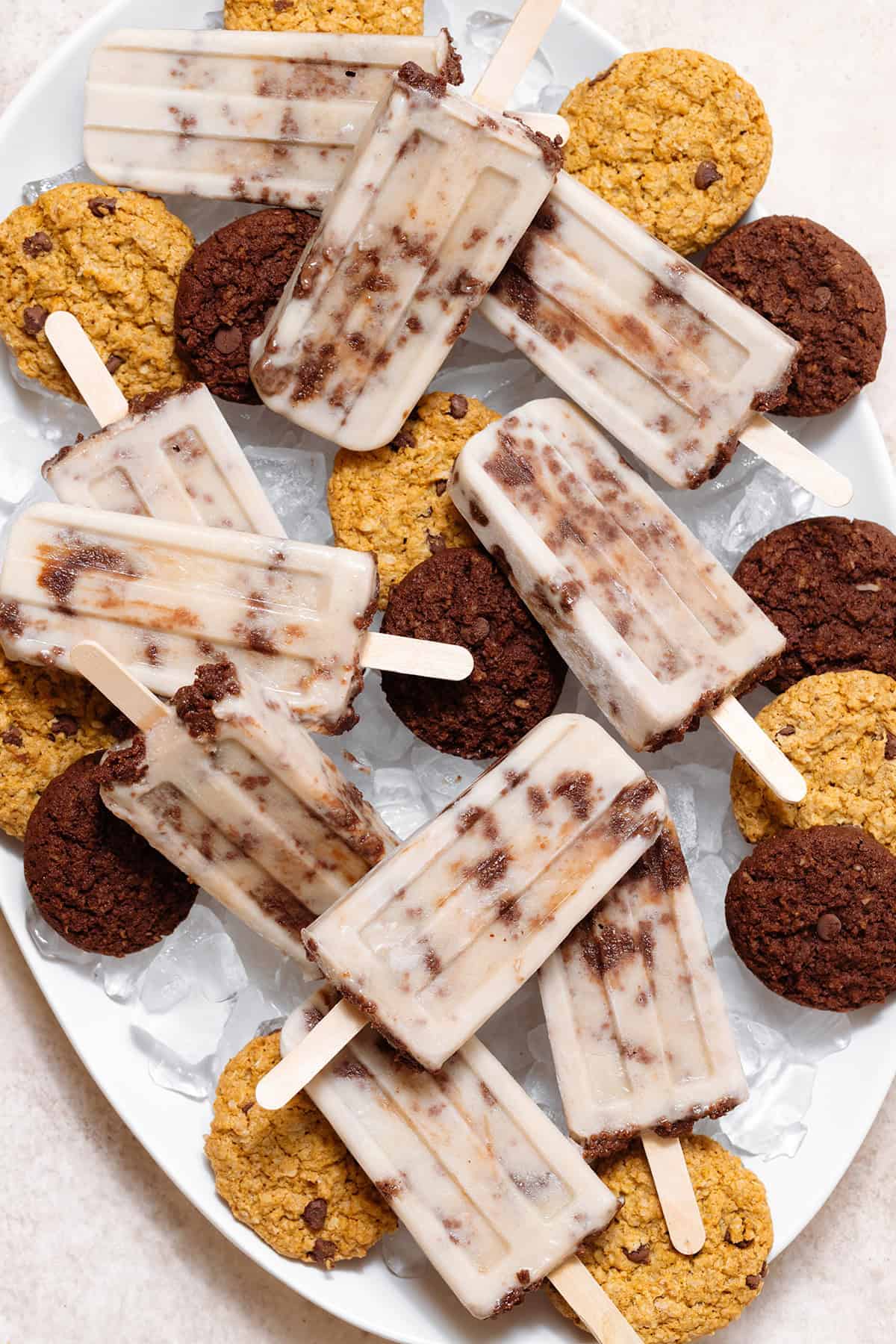 Cookie-dough popsicles arranged on a white serving platter filled with ice and chocolate cookies.