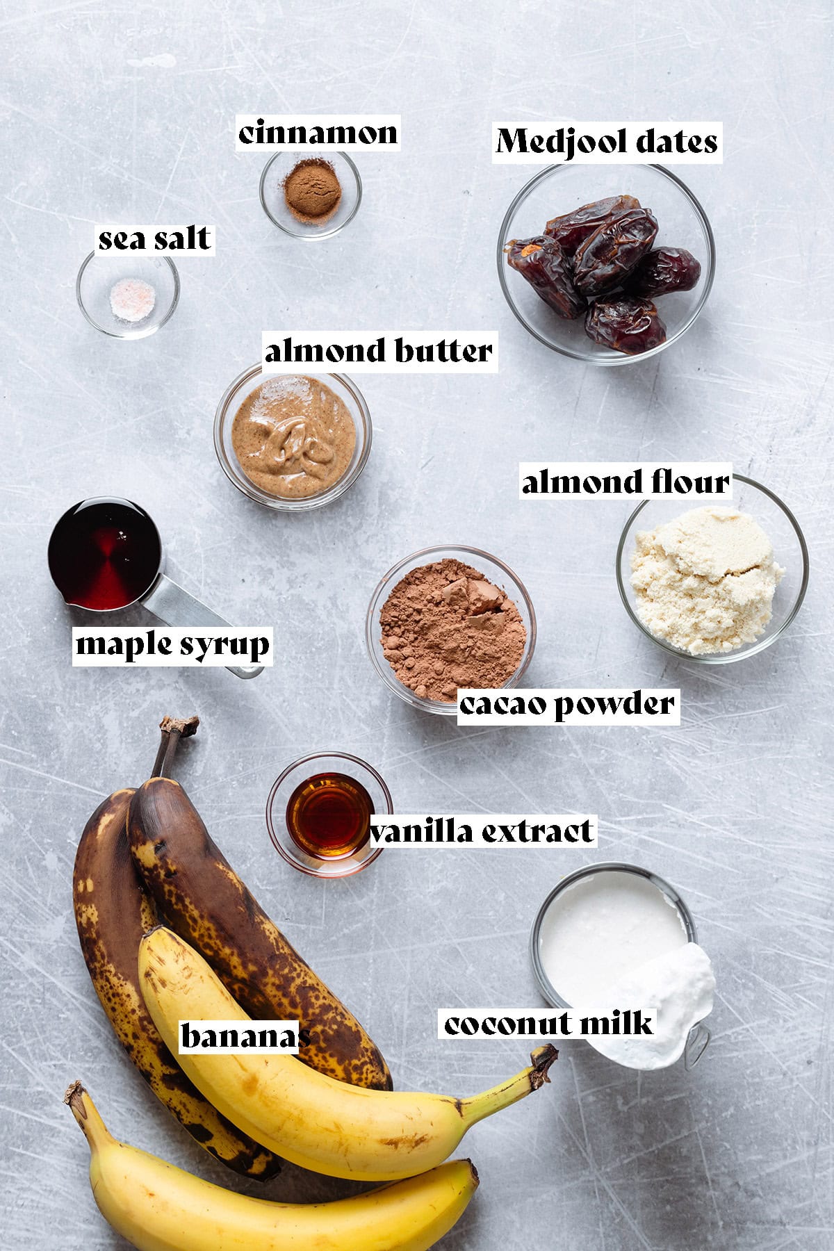 Ingredients for cookie-dough popsicles like ripe bananas, cacao, and almond flour all laid out on a metal background.