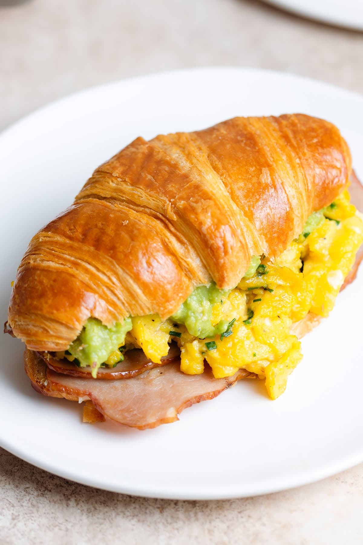 A toasted croissant filled with scrambled eggs, turkey bacon, and avocado on a white plate.