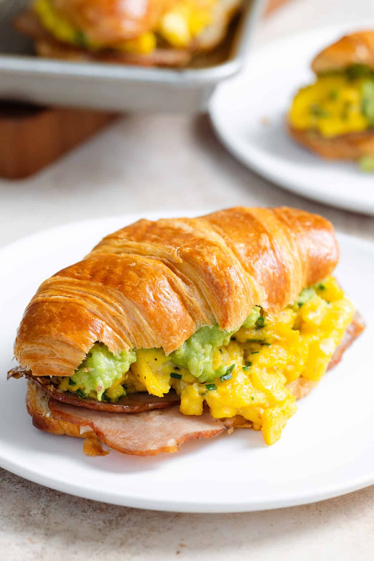 A toasted croissant filled with scrambled eggs, turkey bacon, and avocado on a white plate with more croissants in the background.