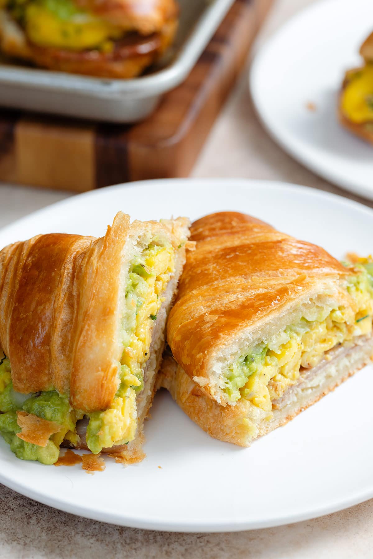 A toasted croissant filled with scrambled eggs, turkey bacon, and avocado that's been cut in half to show what's inside it on a white plate with more croissants in the background.