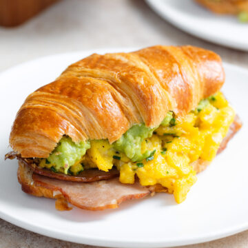 A toasted croissant filled with scrambled eggs, turkey bacon, and avocado on a white plate with more croissants in the background.