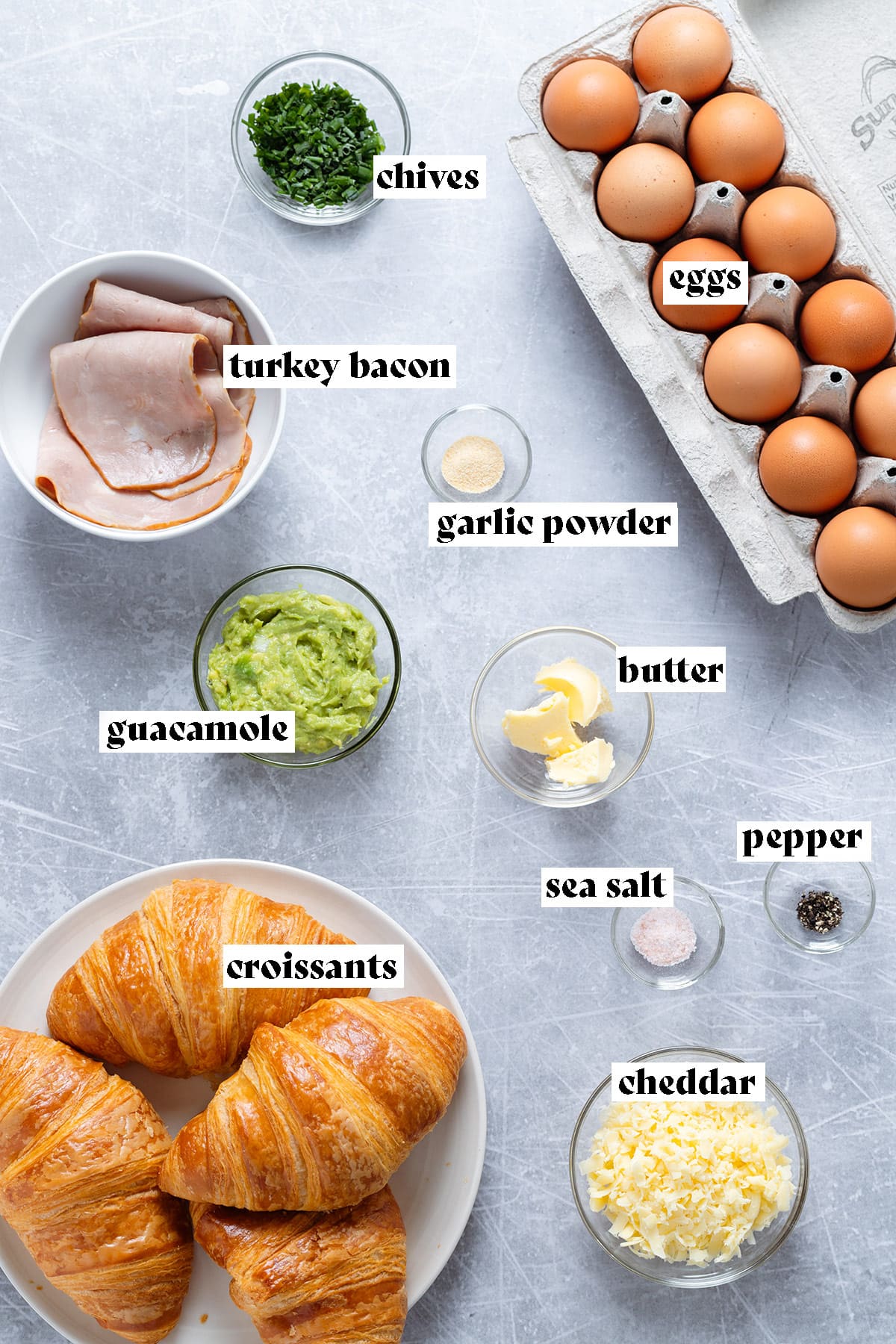 Ingredients for croissant breakfast sandwich like eggs, turkey bacon, and croissants all laid out on a metal background.