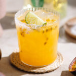 Bright yellow passion fruit margarita in a short salt-rimmed glass with ice and garnished with a slice of lime.